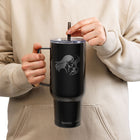 A person in a beige hoodie holds a black insulated tumbler with a handle and clear lid, showing a gray graphic of a woman with flowing hair and a small logo near the bottom. The person is adjusting the black straw.