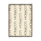 A rectangular woven throw blanket featuring a cream background with repeating vertical 'MONACO' text in dark navy, plus small 'Venezia' details and fringed edges, shown on a plain white background.