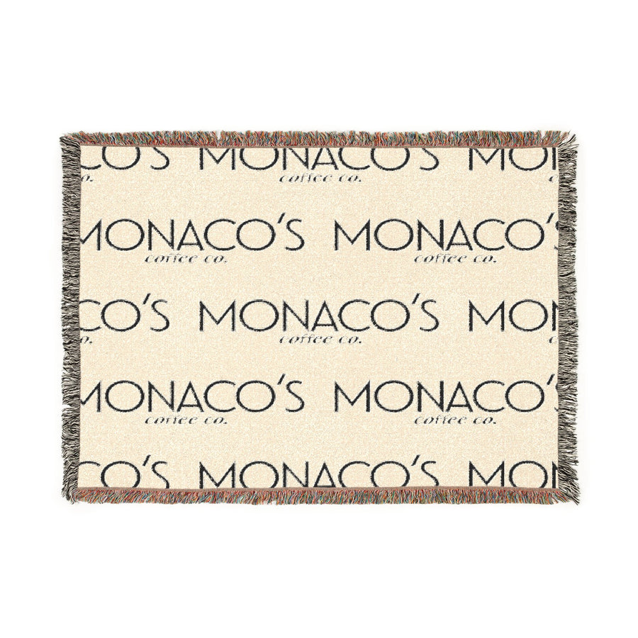 A rectangular woven throw blanket with fringed edges, featuring a cream background with a repeating dark gray 'MONACO'S MOON coffee co.' logo pattern and multicolor fringe.